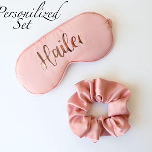 May include: A personalized sleep set in a soft pink hue, featuring a sleep mask with the name "Hailee" cut out and a matching scrunchie. The words "Personalized Set" are written in cursive.