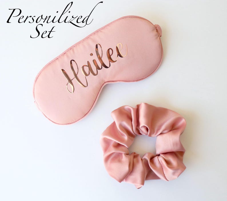 May include: A personalized sleep set in a soft pink hue, featuring a sleep mask with the name "Hailee" cut out and a matching scrunchie. The words "Personalized Set" are written in cursive.