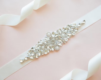 May include: A white satin ribbon with a large, ornate rhinestone embellishment. The embellishment is made of clear rhinestones and pearls, arranged in a floral pattern.