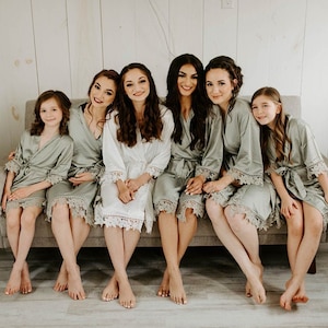May include: Six people, including two young girls, are wearing matching sage green satin robes with white lace trim. They are sitting on a sofa and smiling at the camera.