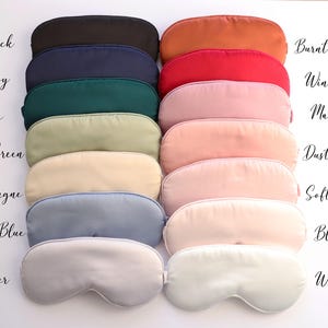 May include: A collection of sleep masks in various colors, including black, navy, and burnt orange. The image displays the masks in a neat arrangement, with each mask showcasing a different shade. Text labels identify each color, such as "Wine Red" and "Dusty Rose."