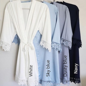 Op de afbeelding: Vijf badjassen in verschillende kleuren: wit, hemelsblauw, stofvrij blauw en marineblauw. Elke badjas heeft een kantversiering en een strikband. De badjassen zijn van links naar rechts gerangschikt in volgorde van kleur.