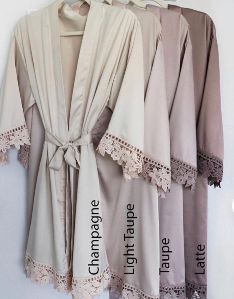 Op de afbeelding: Vier satijnen badjassen in verschillende beige tinten, van de lichtste tot de donkerste: Champagne, Licht Taupe, Taupe en Latte. Elke badjas heeft een delicate kantversiering rond de mouwen en zoom.