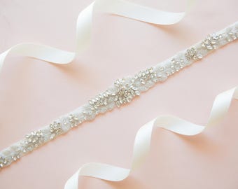 May include: A white satin ribbon with a beaded embellishment. The embellishment is made of clear rhinestones and is arranged in a floral pattern.