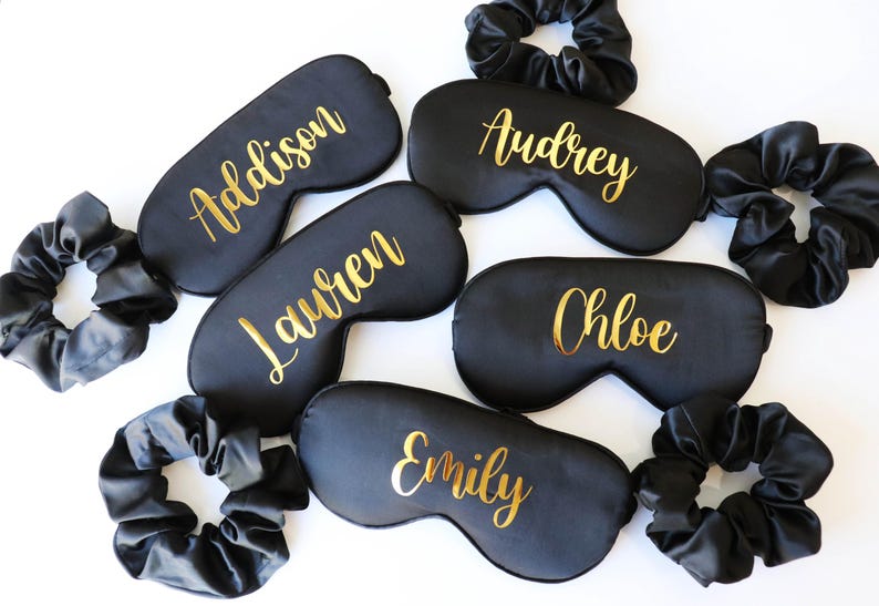 May include: Black sleep masks with gold lettering, each bearing a different name: Addison, Audrey, Lauren, Chloe, and Emily. Matching black scrunchies are also included. These items are likely for a bridal party or a group gift.