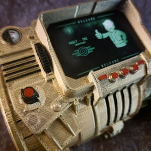 May include: A 3D printed replica of a Pip-Boy from the Fallout video game series. The Pip-Boy is a wearable computer with a screen that displays the text "WELCOME" and "VAULT-TEC" with a Vault-Tec logo. The Pip-Boy has a red button and several other buttons.