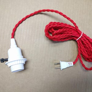 May include: A white light bulb socket with a black turn knob switch attached to a long red fabric-covered twisted electrical cord. The cord is coiled and features a white two-prong electrical plug at the end, secured with a small white tie.