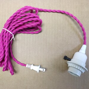 May include: A bright pink fabric-covered twisted electrical cord featuring a white two-prong plug at one end and a white light bulb socket with a small black turn-knob switch at the other end, secured with a white plastic tie.