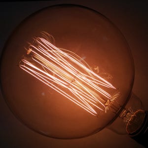 May include: Close-up of a large, round Edison light bulb. The bulb's filament glows with a warm, orange light, contrasting against the clear glass. The bulb's base is black. The overall aesthetic is vintage and industrial.