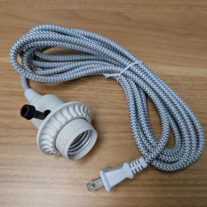 May include: A white and gray fabric-covered electrical cord with a matching light socket and a white plug. The cord is coiled and lies on a wooden surface. The light socket has a black knob.