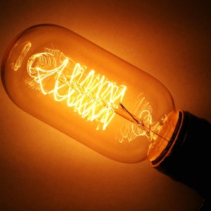 May include: Close-up of a vintage Edison-style light bulb. The bulb has a clear, elongated glass enclosure with a warm, amber glow. The filament is a complex, swirling design, and the base is black.