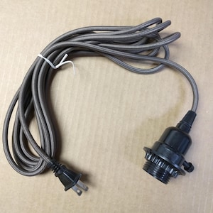 May include: A gray fabric-covered electrical cord with a black lamp socket and a black plug. The cord is bundled and secured with a white tie. The lamp socket has a threaded base for attaching a light bulb.