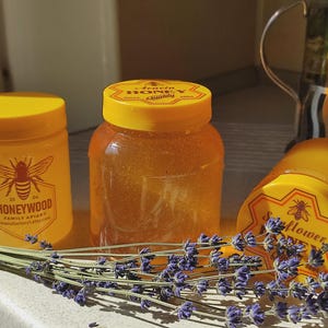 May include: Three clear plastic jars filled with golden honey, each topped with a bright yellow lid. The jars feature labels with bee illustrations and text including Honeywood Family Apiary. A bundle of dried purple lavender stems lies in front.