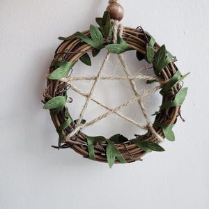 May include: A circular wreath woven from brown twigs, featuring a twine pentagram and green leaf accents. It hangs from a natural cord with a wooden bead. The wreath is a decorative item, suitable for hanging.
