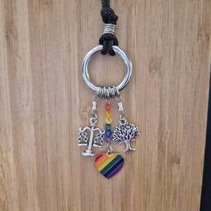May include: A silver-toned metal ring pendant on a black cord featuring three hanging charms: a silver scale of justice, a silver tree, and a central vertical strand of rainbow-coloured beads ending in a heart-shaped pendant with rainbow stripes.