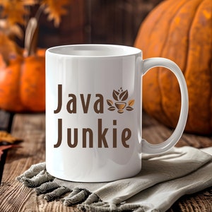 May include: White ceramic coffee mug with a brown graphic that says "Java Junkie" and a small coffee cup with leaves design. The mug is sitting on a wooden table with a white cloth and a pumpkin in the background.