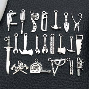 Op de afbeelding: Een verzameling zilverkleurige gereedschap bedels, waaronder een boor, schroevendraaier, rolmaat, zaag, hamer, schop, tang, mes, sleutel, liniaal, bijl en meer. Ideaal voor het maken van sieraden of knutselprojecten.