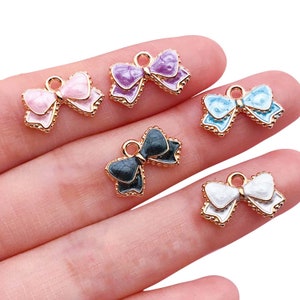 Könnte beinhalten: Fünf goldfarbene Schleifen-Anhänger mit Emaille-Oberflächen in Rosa, Lila, Blau, Schwarz und Weiß. Die Anhänger befinden sich auf einer Hand.