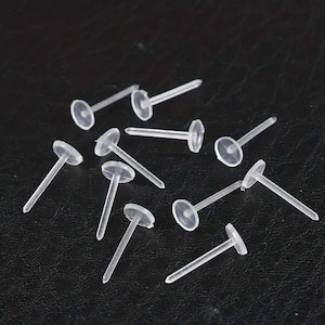 May include: A selection of clear, plastic earring posts. Each post has a flat, circular back and a pointed tip. The posts are scattered on a dark surface, highlighting their simple design and transparency. Ideal for jewellery making.