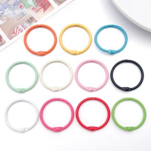 May include: A collection of colorful binder rings in various shades, including orange, yellow, blue, green, white, pink, and black. Each ring has a small, matching clasp for easy opening and closing. These rings are ideal for organizing papers or crafts.