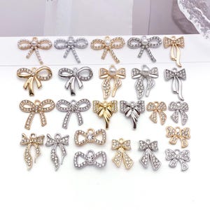 May include: A collection of twenty-four metal bow-shaped charms arranged in five rows. The charms feature gold and silver tones, embellished with clear rhinestones or small white faux pearls. Each piece includes a small loop for attachment.