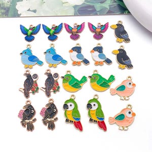 Puede incluir: Una colección de diecinueve dijes de metal con forma de pájaro y diseños de esmalte de colores, que incluyen colibríes, arrendajos azules, cuervos sobre ramas con flores, loros verdes y pequeños pájaros rosas, cada uno con una argolla.