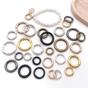 Può includere: Una collezione di anelli con chiusura a molla in metallo di vari colori: oro, argento, nero e bronzo. Sono presenti anche un braccialetto di perle e un charm. Queste chiusure sono adatte per la creazione di gioielli e l'artigianato.