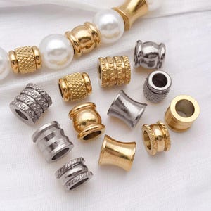 May include: A collection of gold and silver-coloured metal beads in diverse shapes and textures. Some are cylindrical, others barrel-shaped, and some have textured surfaces. A strand of white pearl beads is visible in the background.