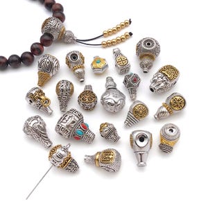 Op de afbeelding: Een verzameling versierde metalen kralen met ingewikkelde zilver- en goudkleurige patronen, sommige ingelegd met kleine turquoise en rode stenen. De kralen variëren in vorm, waaronder kalebasachtige en geometrische ontwerpen.