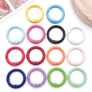 Op de afbeelding: Een verzameling kleurrijke, ronde karabijnhaakclips in diverse tinten, waaronder geel, blauw, roze, zwart, wit, rood, oranje en groen. Deze metalen clips hebben een veergrendel voor veilige bevestiging en zijn ideaal voor sleutelhangers of knutselprojecten.