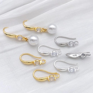 May include: An assortment of gold and silver-toned earring findings. Some feature a curved hook design with a small, clear gemstone accent. Other findings have a pearl drop or a small loop for attaching a charm. The background is a white, textured fabric.