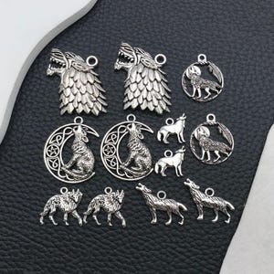 May include: A collection of silver-toned metal charms featuring wolf motifs. The set includes two wolf head charms, two wolf-in-crescent-moon charms, two circular wolf charms, two small standing wolf charms, and four walking wolf charms with loops.