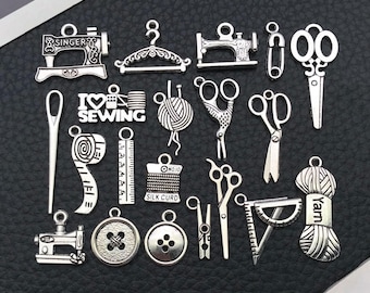 Puede incluir: Colección de dijes de costura plateados, que incluyen máquinas de coser, tijeras, botones, cintas métricas y ovillos de hilo. Algunos dijes tienen las palabras "I Love Sewing" y "Singer". Los dijes están dispuestos sobre una superficie oscura.