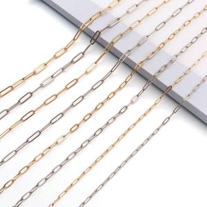 May include: Close-up of several paperclip-style chain necklaces in gold and silver tones. The chains are arranged diagonally across the frame, showcasing their elongated oval links. The background is a clean, white surface.