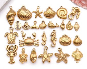Puede incluir: Una colección de dijes dorados con temática marina, que incluye estrellas de mar, conchas, caballitos de mar y un cangrejo. Cada dije tiene un pequeño lazo para sujetarlo a joyas u otras manualidades. Los dijes están dispuestos sobre una superficie blanca.