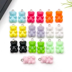 Könnte beinhalten: Eine Sammlung bunter Gummibärchen-Anhänger in verschiedenen Farben, darunter Blau, Rot, Orange, Gelb, Lila, Weiß, Schwarz, Grün und Rosa. Jeder Anhänger hat eine silberne Schlaufe zum Befestigen an Schmuck oder Bastelarbeiten.