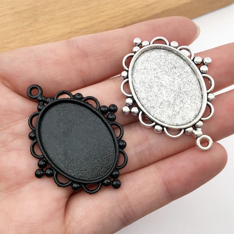 May include: Two oval-shaped metal pendant trays with ornate edges. One tray is silver with a smooth surface and the other is black with a textured surface.