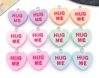 May include: A collection of twelve heart-shaped charms in various pastel colors, each with a silver loop and the words "HUG ME" in red. The charms are in shades of lavender, peach, pink, and mint green.