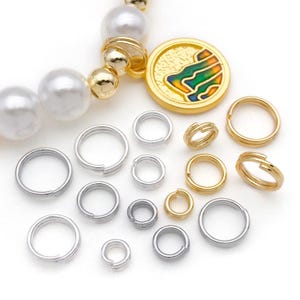 May include: An assortment of jewelry-making supplies, including silver and gold-colored split rings, a gold pendant with colorful enamel, and a beaded strand with white and gold beads. The items are arranged on a white surface.