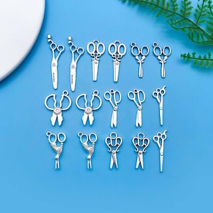 May include: A collection of 15 silver-toned metal scissor charms. The charms are in various styles, including traditional scissors, decorative scissors, and scissors with a bird design. The charms are perfect for adding a touch of whimsy to jewellery or crafts.