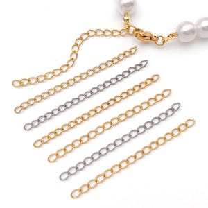 Puede incluir: Primer plano de extensores de cadena dorados y plateados para joyería. Los extensores son de varias longitudes y están conectados a un cierre dorado y un collar de perlas. El fondo es blanco.