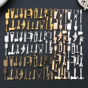 Pode incluir: Uma coleção de berloques de metal com formas de diversas ferramentas manuais, incluindo serras, martelos, chaves inglesas, chaves de fenda e machados. Os berloques têm acabamentos em tons de ouro, prata e bronze dispostos em uma grade.