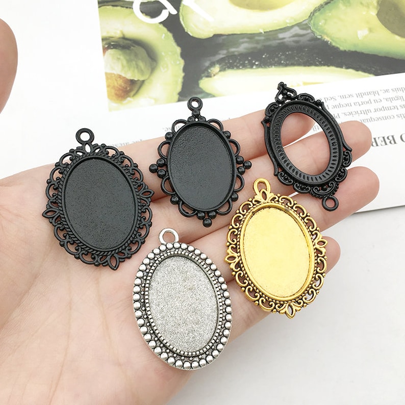 May include: Four oval-shaped metal pendant trays with different finishes: black, gold, and silver. The trays have intricate designs around the edges and are ready for crafting projects.