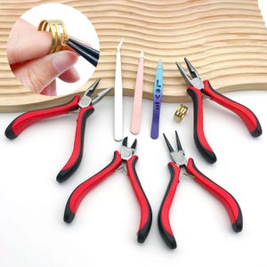May include: A selection of jewellery-making tools, including red and black pliers, tweezers, and a ring. The pliers have red handles and black grips. The tweezers are white, pink, and blue. The ring is gold-coloured. The tools are arranged on a wooden surface.