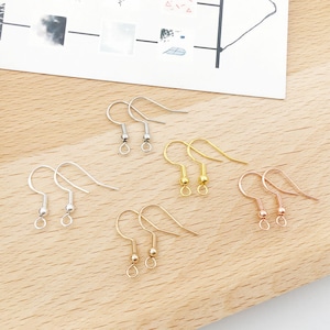 May include: An assortment of earring hooks in silver, gold, and rose gold tones. The hooks are displayed on a light wooden surface, with a piece of paper in the background. The hooks are designed for jewelry making and crafting.