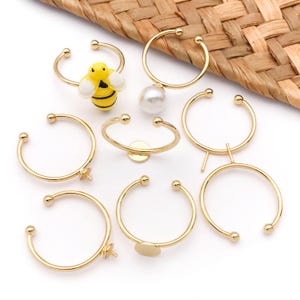 Könnte beinhalten: Eine Sammlung goldfarbener Ringrohlinge mit verschiedenen dekorativen Elementen. Einige Ringe haben einen kleinen gelb-schwarzen Bienenanhänger, eine weiße Perle oder eine flache goldene Scheibe. Die Ringe sind auf einer weißen Oberfläche mit gewebtem Hintergrund angeordnet.