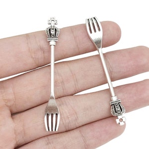 May include: Two silver-toned forks with crown-shaped tops. The forks are held in a hand.