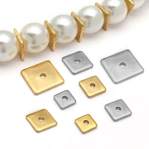 May include: A collection of square metal beads in gold and silver tones, each featuring a central hole. The assortment includes various sizes of flat, square spacers arranged alongside a strand of white spherical pearls separated by gold square spacers.