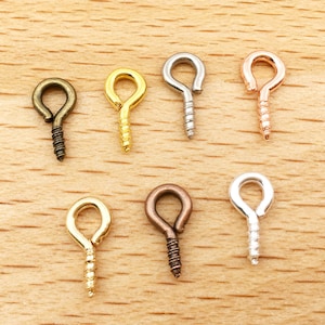 May include: A collection of seven metal screw eyelets in various finishes: antique bronze, gold, silver, and rose gold. The eyelets are arranged on a light wood surface, showcasing their screw threads and looped tops, ideal for jewelry making or crafts.