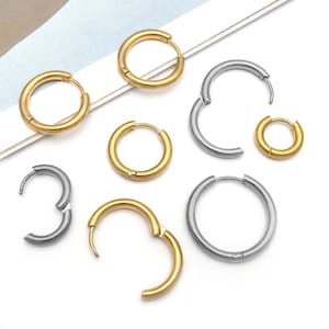 May include: An assortment of gold and silver hoop earrings in various sizes. Some hoops are closed, while others are open, showcasing the clasp mechanism. The earrings are arranged on a white surface, highlighting their metallic shine and simple design.